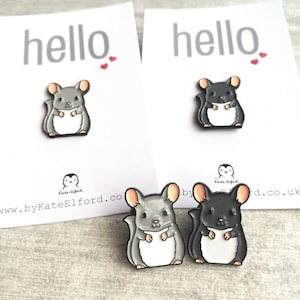 May include: Two enamel pins featuring a cartoon chinchilla. The chinchilla on the left is grey and the chinchilla on the right is black. Both chinchillas are standing on their hind legs with their front paws raised. The pins are on a white background with the word "hello" printed on the top of each card.