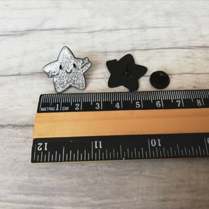 May include: Two star-shaped enamel pins. One is silver glitter with a smiling face and outstretched arms. The other is black, showing the back with a pin clasp. A ruler shows measurements in centimetres.