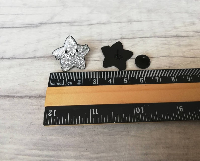 May include: Two star-shaped enamel pins. One is silver glitter with a smiling face and outstretched arms. The other is black, showing the back with a pin clasp. A ruler shows measurements in centimetres.