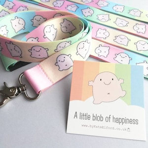 May include: A pastel-coloured lanyard featuring a repeating pattern of smiling blob characters. The lanyard has a silver-coloured clasp and a matching card that reads "A little blob of happiness". The lanyard is approximately 91 cm long.