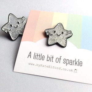 May include: Two silver glitter star-shaped pins with black outlines. The stars have smiling faces and outstretched arms. The pins are displayed on a card with the text "A little bit of sparkle" and a rainbow background.