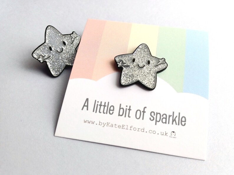 May include: Two silver glitter star-shaped pins with black outlines. The stars have smiling faces and outstretched arms. The pins are displayed on a card with the text "A little bit of sparkle" and a rainbow background.