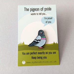 A colourful pigeon pin on a card. The pin has a light blue body, black wings, and pink details. The card reads "The pigeon of pride" and "I'm proud of you."