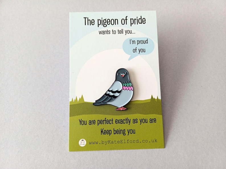 A colourful pigeon pin on a card. The pin has a light blue body, black wings, and pink details. The card reads "The pigeon of pride" and "I'm proud of you."