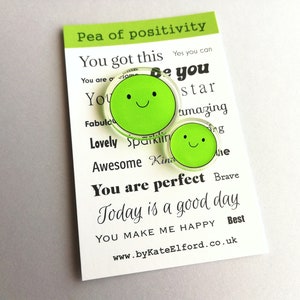 Puede incluir: Dos pines verdes con forma de guisante y caras sonrientes, presentados en una tarjeta blanca. La tarjeta muestra "Pea of positivity" y frases inspiradoras como "You got this" y "You are perfect."
