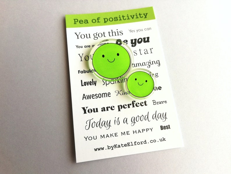 Puede incluir: Dos pines verdes con forma de guisante y caras sonrientes, presentados en una tarjeta blanca. La tarjeta muestra "Pea of positivity" y frases inspiradoras como "You got this" y "You are perfect."