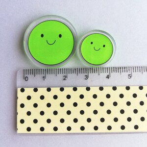 Puede incluir: Dos c&iacute;rculos transparentes de acr&iacute;lico con un dise&ntilde;o de cara verde sonriente. El c&iacute;rculo m&aacute;s grande mide aproximadamente 5 cm de di&aacute;metro, y el m&aacute;s peque&ntilde;o, unos 2,5 cm. Una regla con medidas en cm est&aacute; colocada junto a los c&iacute;rculos.