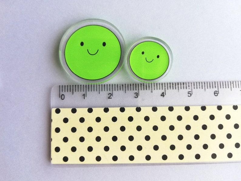 Puede incluir: Dos c&iacute;rculos transparentes de acr&iacute;lico con un dise&ntilde;o de cara verde sonriente. El c&iacute;rculo m&aacute;s grande mide aproximadamente 5 cm de di&aacute;metro, y el m&aacute;s peque&ntilde;o, unos 2,5 cm. Una regla con medidas en cm est&aacute; colocada junto a los c&iacute;rculos.