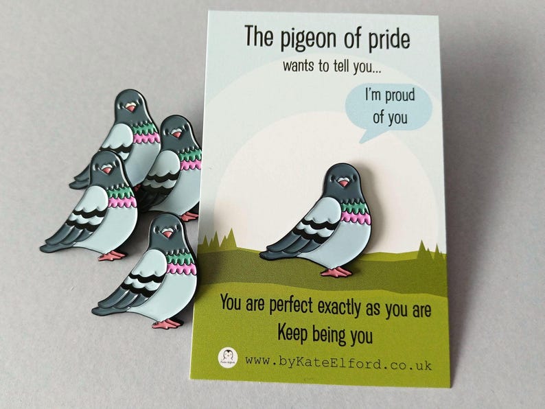 A set of pigeon enamel pins in shades of grey, light blue, pink, and green, alongside a card. The card reads "The pigeon of pride wants to tell you... I'm proud of you. You are perfect exactly as you are. Keep being you."