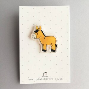 May include: A yellow donkey with a black mane and hooves, illustrated on a clear acrylic pin. The pin is on a white card with black polka dots and the text "www.byKateElford.co.uk".