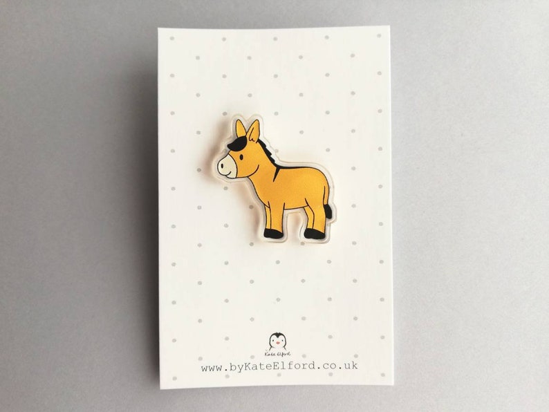 May include: A yellow donkey with a black mane and hooves, illustrated on a clear acrylic pin. The pin is on a white card with black polka dots and the text "www.byKateElford.co.uk".