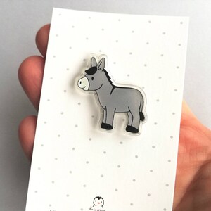 May include: A gray donkey cartoon character on a clear acrylic pin. The pin is on a white card with small gray dots.