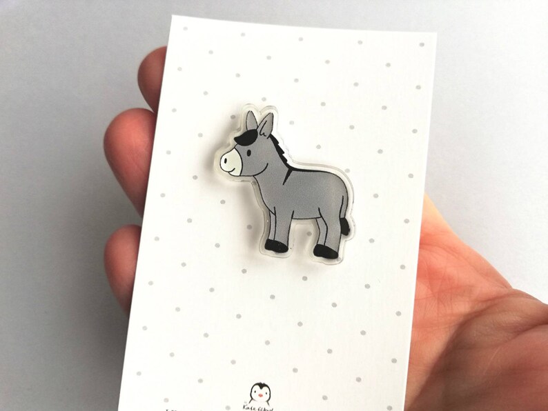 May include: A gray donkey cartoon character on a clear acrylic pin. The pin is on a white card with small gray dots.