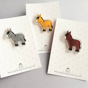 May include: Three donkey pins, each with a different color: gray, brown, and yellow. The pins are made of clear acrylic and have a black outline. The pins are displayed on white cards with black polka dots and the website address "www.byKateElford.co.uk".