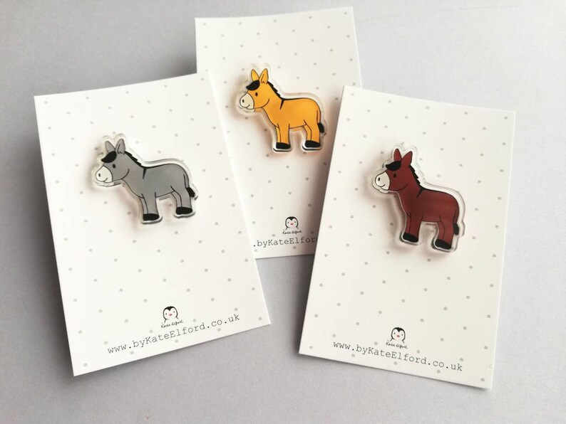 May include: Three donkey pins, each with a different color: gray, brown, and yellow. The pins are made of clear acrylic and have a black outline. The pins are displayed on white cards with black polka dots and the website address "www.byKateElford.co.uk".