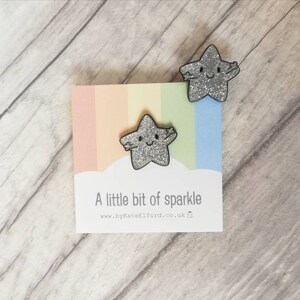 May include: Two glittery silver star-shaped pins with smiling faces and outstretched arms are displayed on a card with a pastel rainbow background. The card reads "A little bit of sparkle" and the website address.