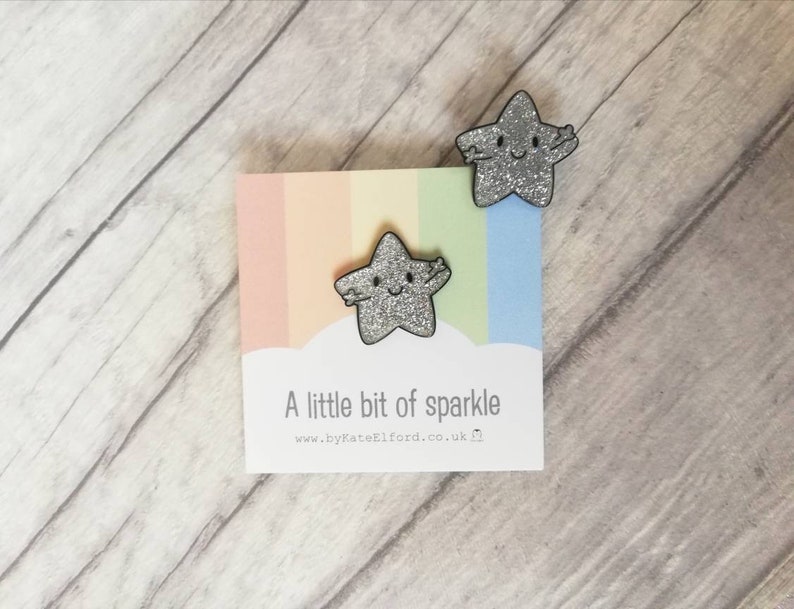 May include: Two glittery silver star-shaped pins with smiling faces and outstretched arms are displayed on a card with a pastel rainbow background. The card reads "A little bit of sparkle" and the website address.