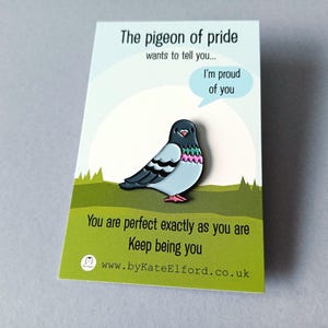 A pin badge depicting a cartoon pigeon with a light blue body, black wings, and pink and green details. The badge is on a card with the text "The pigeon of pride wants to tell you... I'm proud of you." and "You are perfect exactly as you are. Keep being you."