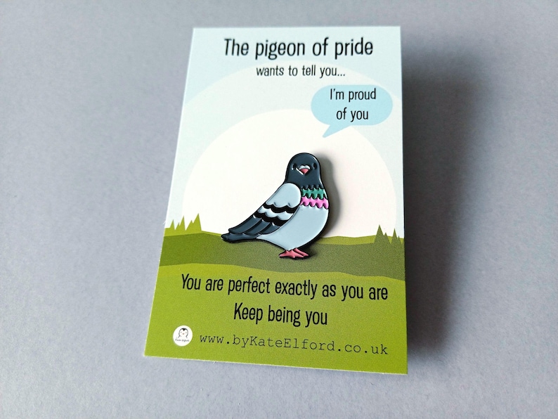 A pin badge depicting a cartoon pigeon with a light blue body, black wings, and pink and green details. The badge is on a card with the text "The pigeon of pride wants to tell you... I'm proud of you." and "You are perfect exactly as you are. Keep being you."