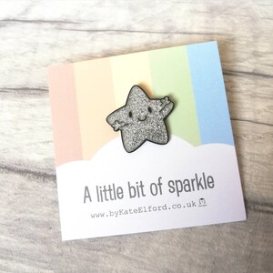 May include: A silver glitter star-shaped pin with a smiling face and outstretched arms, on a card with a rainbow gradient and the text "A little bit of sparkle". A charming, whimsical accessory.