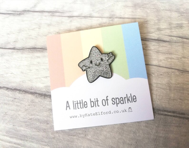 May include: A silver glitter star-shaped pin with a smiling face and outstretched arms, on a card with a rainbow gradient and the text "A little bit of sparkle". A charming, whimsical accessory.