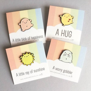 Four enamel pins with colourful backgrounds and rainbow stripes. The pins feature cartoon characters with different expressions and text that reads "A little blob of happiness", "A hug", "A little ray of sunshine", and "A worry gobbler".