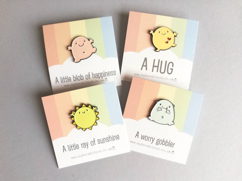 Four enamel pins with colourful backgrounds and rainbow stripes. The pins feature cartoon characters with different expressions and text that reads "A little blob of happiness", "A hug", "A little ray of sunshine", and "A worry gobbler".