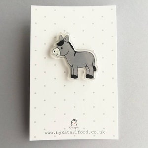 May include: A gray donkey cartoon character on a clear acrylic pin. The pin is on a white background with small gray dots.