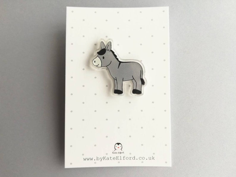 May include: A gray donkey cartoon character on a clear acrylic pin. The pin is on a white background with small gray dots.