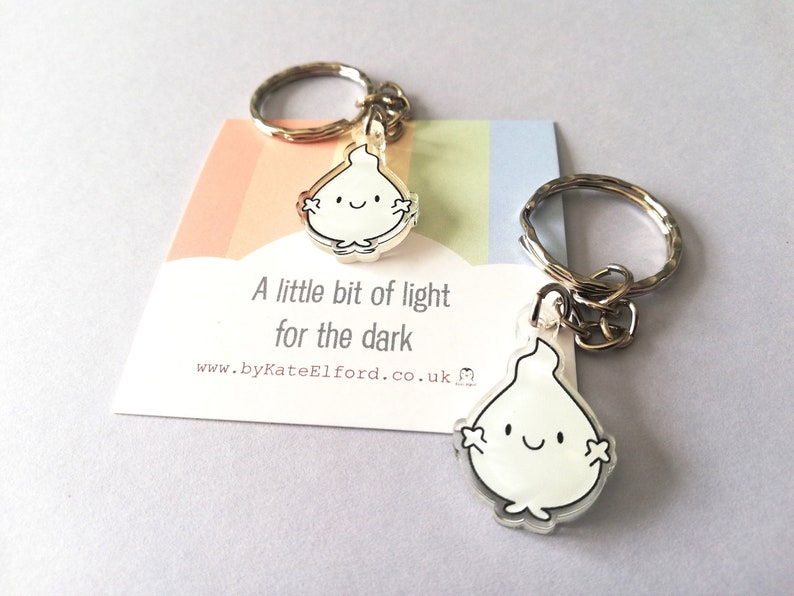 Two silver keyrings with a clear, white flame charm. The flame has a smiling face and small arms. The charms are attached to silver key rings. The keyrings are displayed on a card with the text "A little bit of light for the dark".