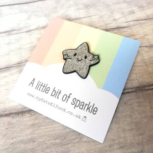 May include: A sparkly silver star-shaped brooch with a black border and a smiling face. The brooch is on a card with a rainbow design and the text "A little bit of sparkle".