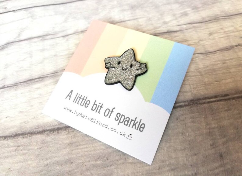 May include: A sparkly silver star-shaped brooch with a black border and a smiling face. The brooch is on a card with a rainbow design and the text "A little bit of sparkle".