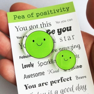 Puede incluir: Dos pines verdes con forma de guisante y caras sonrientes se muestran en una tarjeta blanca con el texto "Pea of positivity". La tarjeta tambi&eacute;n tiene afirmaciones positivas como "You got this" y "You are perfect".