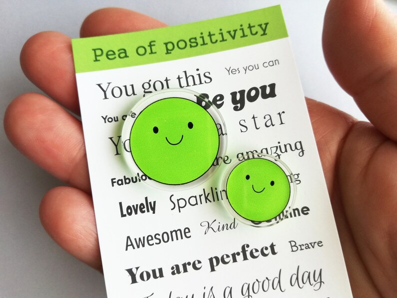 Puede incluir: Dos pines verdes con forma de guisante y caras sonrientes se muestran en una tarjeta blanca con el texto "Pea of positivity". La tarjeta tambi&eacute;n tiene afirmaciones positivas como "You got this" y "You are perfect".