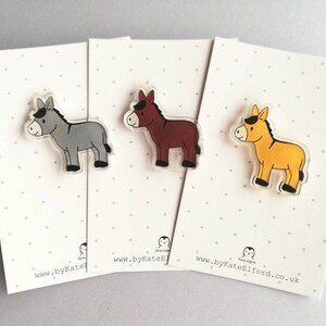 May include: Three donkey pins, each with a different color: gray, brown, and yellow. The pins are on white cards with black polka dots and the website address "www.byKateElford.co.uk".