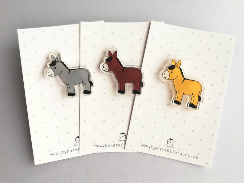 May include: Three donkey pins, each with a different color: gray, brown, and yellow. The pins are on white cards with black polka dots and the website address "www.byKateElford.co.uk".