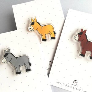 May include: Three donkey pins, one gray, one yellow, and one brown. The pins are made of clear acrylic with a cartoon donkey design.