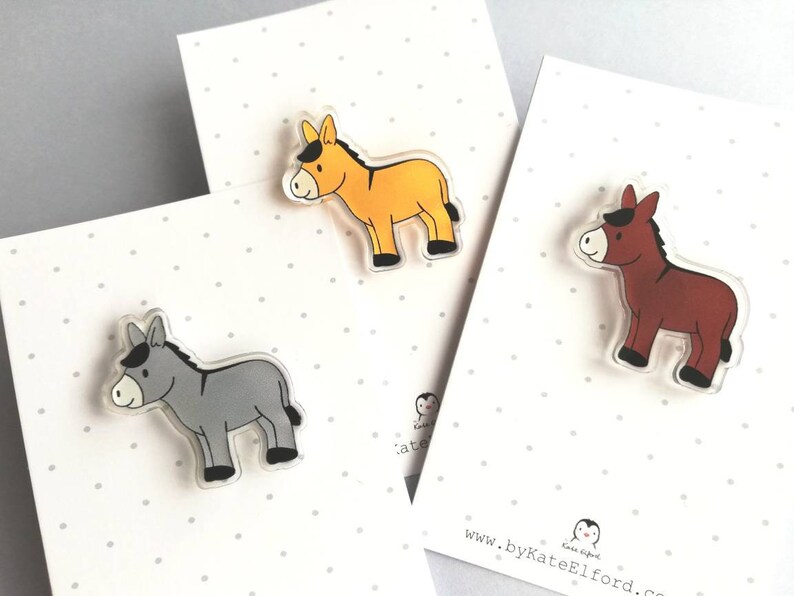 May include: Three donkey pins, one gray, one yellow, and one brown. The pins are made of clear acrylic with a cartoon donkey design.