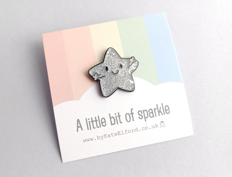 A silver glitter star pin, with a happy face and arms held out for a hug. The backing card has a rainbow background with the wording a little bit of sparkle