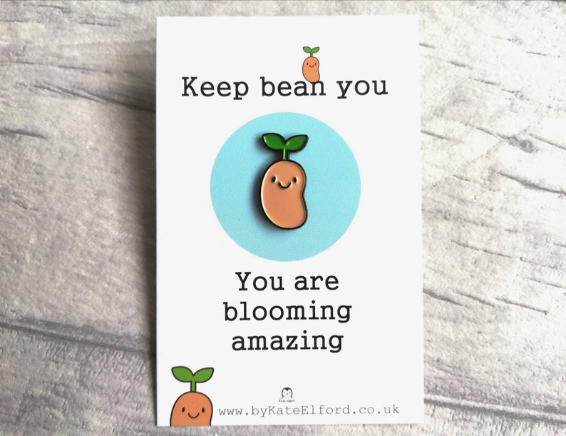 A cute little bean enamel pin. The bean has a green shoot with two little leaves sticking out and a happy face. The backing card says keep bean you. You are blooming amazing. A cute and positive little gift