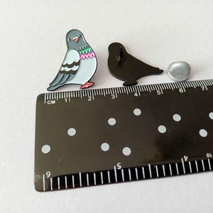 Two pigeon-shaped enamel pins. One pin is a colorful pigeon with grey, blue, pink, and green details. The other is a black silhouette of a pigeon. A ruler shows the pins are approximately 2.5 cm in length.