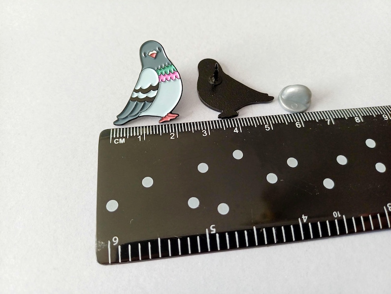 Two pigeon-shaped enamel pins. One pin is a colorful pigeon with grey, blue, pink, and green details. The other is a black silhouette of a pigeon. A ruler shows the pins are approximately 2.5 cm in length.