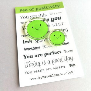 Puede incluir: Dos pines verdes con forma de guisante con caras sonrientes, uno grande y otro peque&ntilde;o, se muestran en una tarjeta blanca. La tarjeta tiene el texto "Pea of positivity" y palabras inspiradoras como "You are perfect" y "Today is a good day."