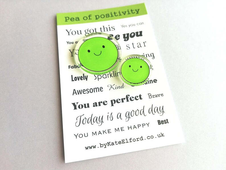 Puede incluir: Dos pines verdes con forma de guisante con caras sonrientes, uno grande y otro peque&ntilde;o, se muestran en una tarjeta blanca. La tarjeta tiene el texto "Pea of positivity" y palabras inspiradoras como "You are perfect" y "Today is a good day."