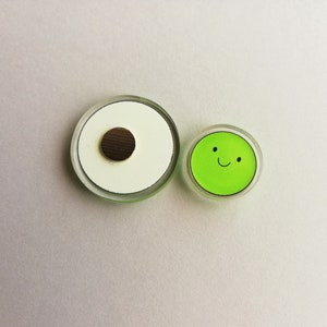 Puede incluir: Dos imanes redondos. Uno es blanco con un centro marr&oacute;n, el otro es verde con una cara sonriente. Ambos est&aacute;n encapsulados en pl&aacute;stico transparente con un borde verde. Imanes para nevera o pizarra.