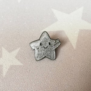 May include: A silver glitter star-shaped pin with a smiling face and outstretched arms. The pin has a black outline. The background is light pink with star shapes.