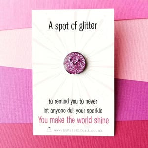 May include: A pink glittery badge with a smiling face, on a white card with text. The card says "A spot of glitter" and "You make the world shine".