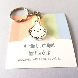 A silver keyring with a small white flame charm. The charm has a happy face and the text "A little bit of light for the dark" is printed on a card attached to the keyring.