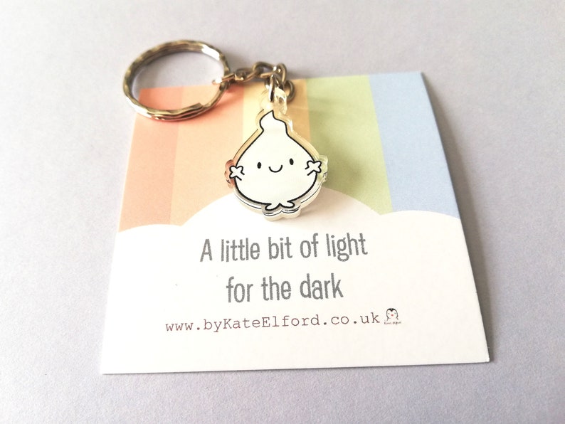 A silver keyring with a small white flame charm. The charm has a happy face and the text "A little bit of light for the dark" is printed on a card attached to the keyring.