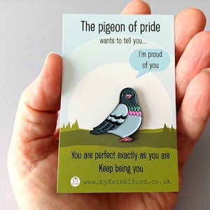 A pin badge depicting a cartoon pigeon with a light blue body, black wings, and pink and green details. The badge is on a card with the text "The pigeon of pride wants to tell you... I'm proud of you." and "You are perfect exactly as you are. Keep being you."
